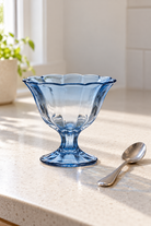 Abby Footed Glass Bowl - Blue on a pale kitchen bench beside a teaspoon, showing translucent glass; Coast to Coast TW1116
