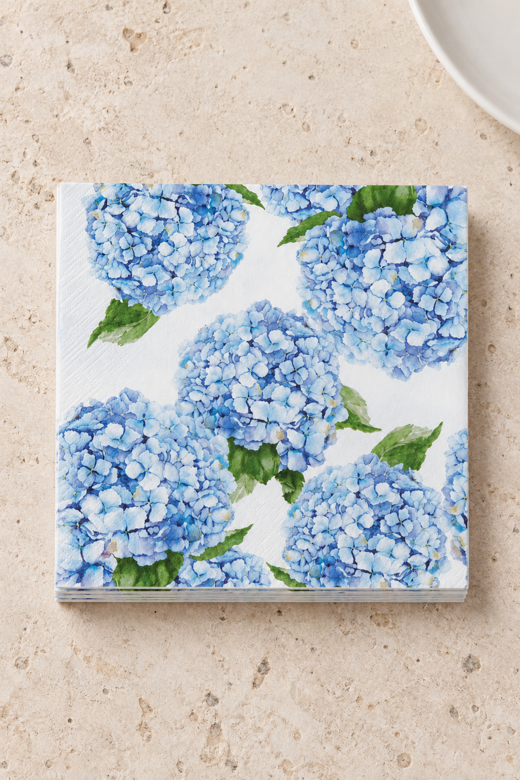 Hydrangea Paper Napkins - Blue in a precisely aligned thin paper stack, viewed overhead on a pale stone bench; Coast to Coast KW5355